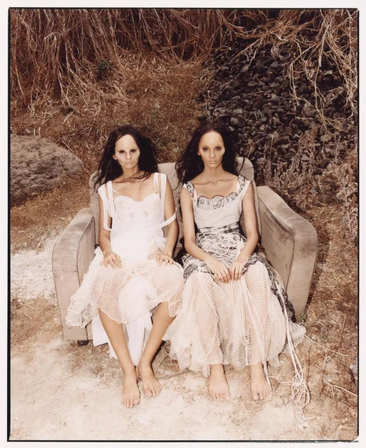 Two women in vintage dresses sit on a worn armchair outdoors against a backdrop of dry grass and foliage in a fine art fashion shot.