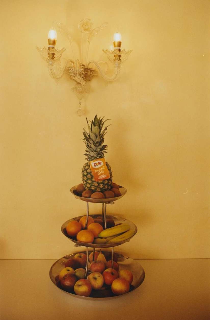 Three-tiered fruit stand featuring a pineapple on top, surrounded by kiwis, oranges, bananas, and apples, set against a yellow background.