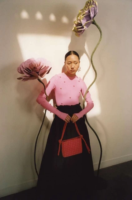 A stylish Asian woman poses indoors with oversized paper flowers, wearing a pink long-sleeve top, black skirt, and holding a red woven bag.