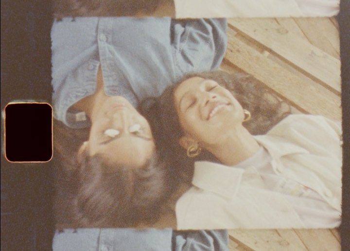 Two women lie side-by-side smiling with curly dark hair on wood slats, wearing casual tops and hoop earrings, lit by daylight.
