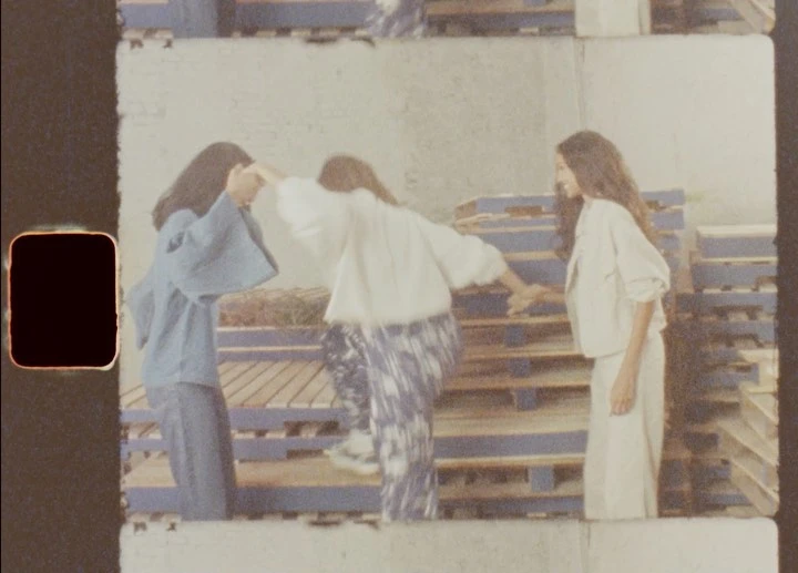 Three women pose together indoors, wearing casual tops and patterned pants, with a backdrop of blue and brown wooden pallets.