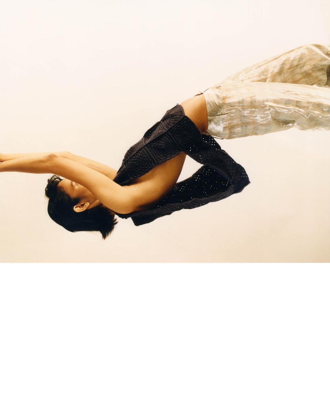 Adult female fashion model poses wearing a dark knitted top and light-colored trousers against a neutral backdrop in a studio setting.
