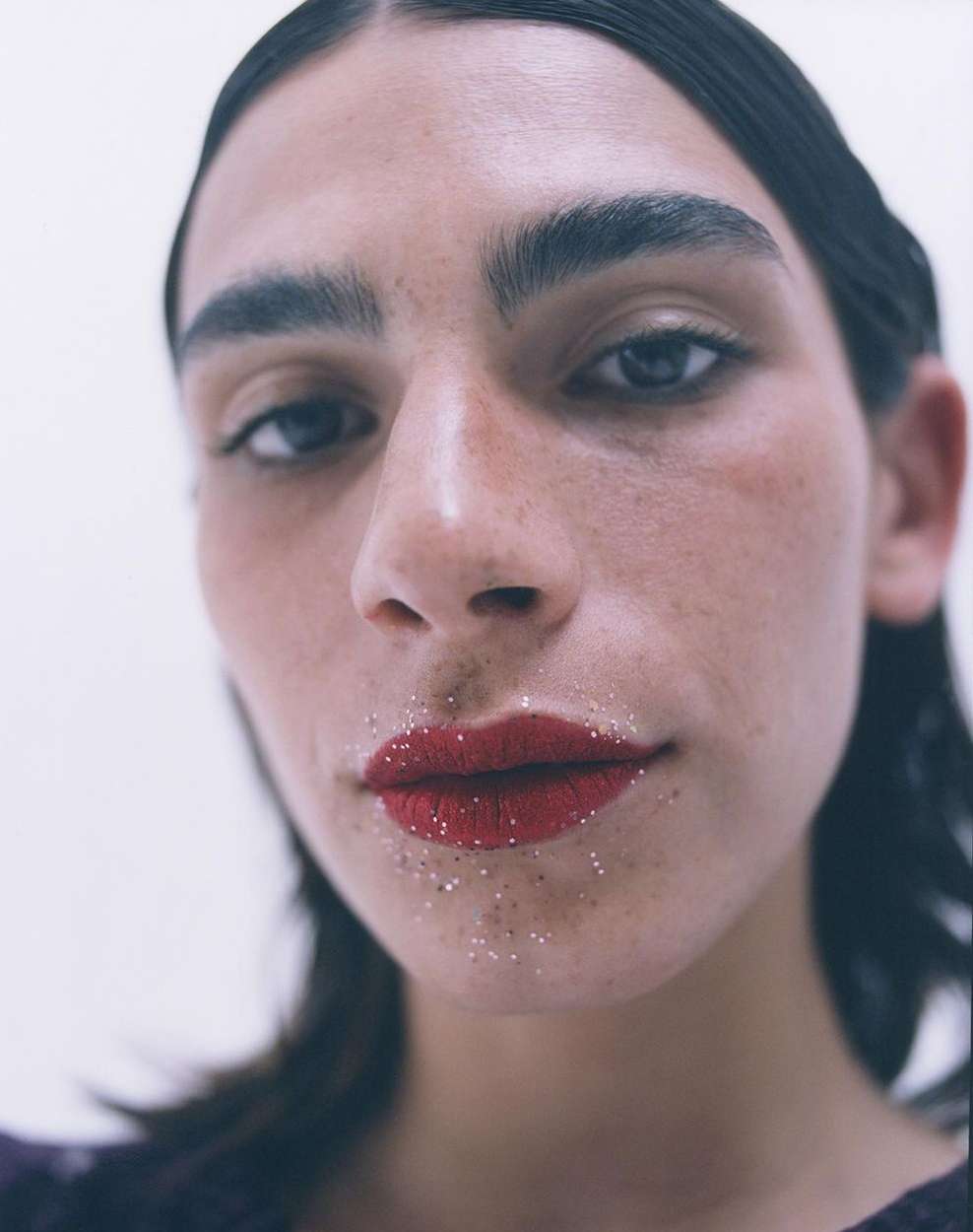 Close-up of an adult female with red lipstick and glitter, gazing upward against a plain white background in a beauty editorial shot.