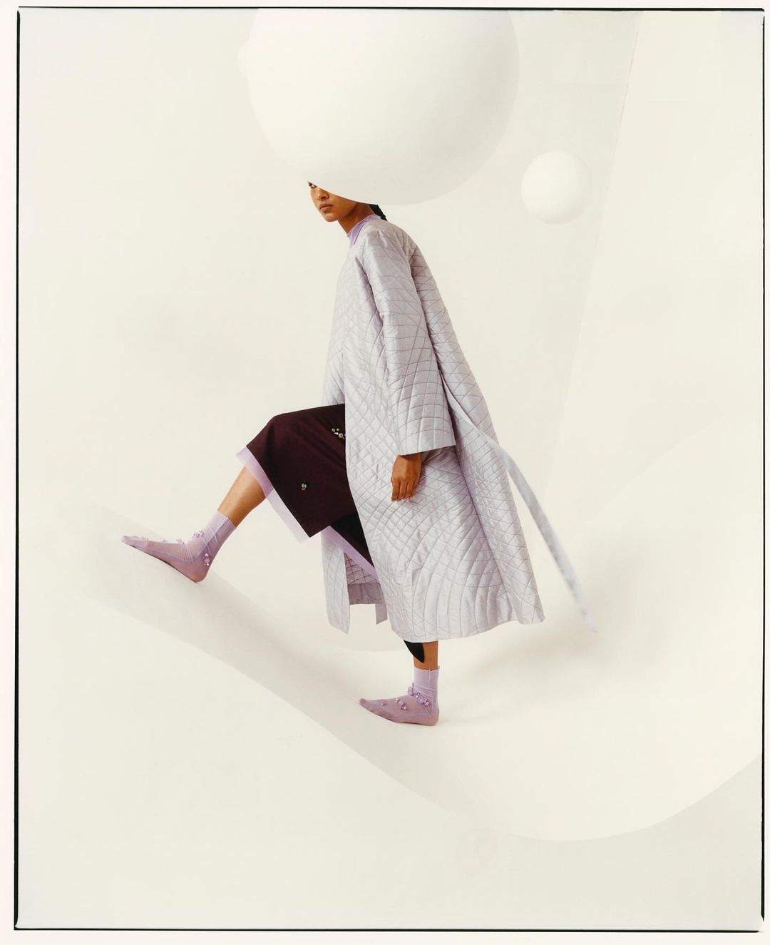 Fashion editorial shot of a woman in a long, light purple coat and socks, stepping down a cream-colored surface with large, white spheres.