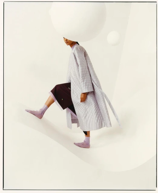 Fashion editorial shot of a woman in a long, light purple coat and socks, stepping down a cream-colored surface with large, white spheres.