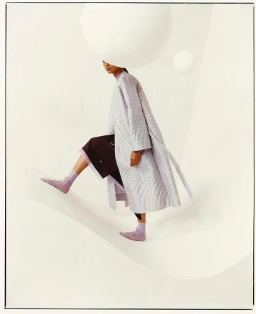 Fashion editorial shot of a woman in a long, light purple coat and socks, stepping down a cream-colored surface with large, white spheres.