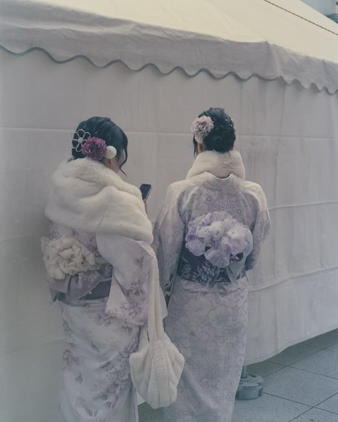 Two women in kimonos stand closely together, backs to the camera, adorned with floral hairpieces and fur stoles against a plain white backdrop.