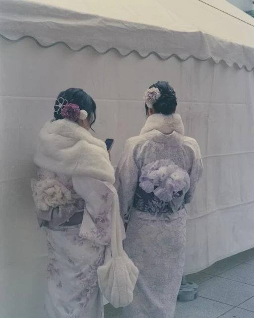 Two women in kimonos stand closely together, backs to the camera, adorned with floral hairpieces and fur stoles against a plain white backdrop.