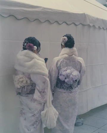 Two women in kimonos stand closely together, backs to the camera, adorned with floral hairpieces and fur stoles against a plain white backdrop.