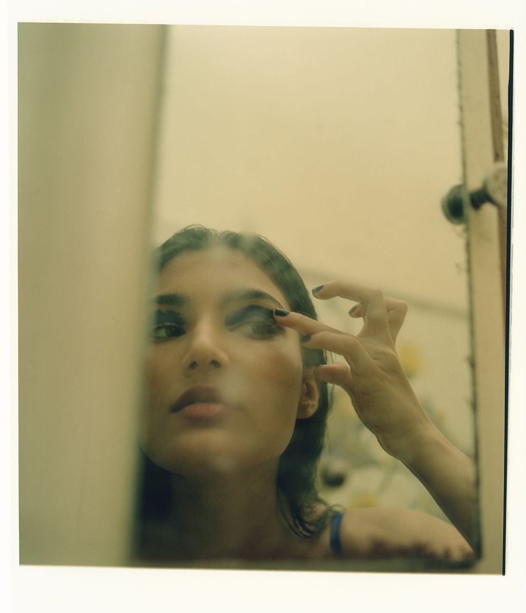 Young woman applying dark eye shadow while looking in a mirror with an off-white frame in an indoor environment.