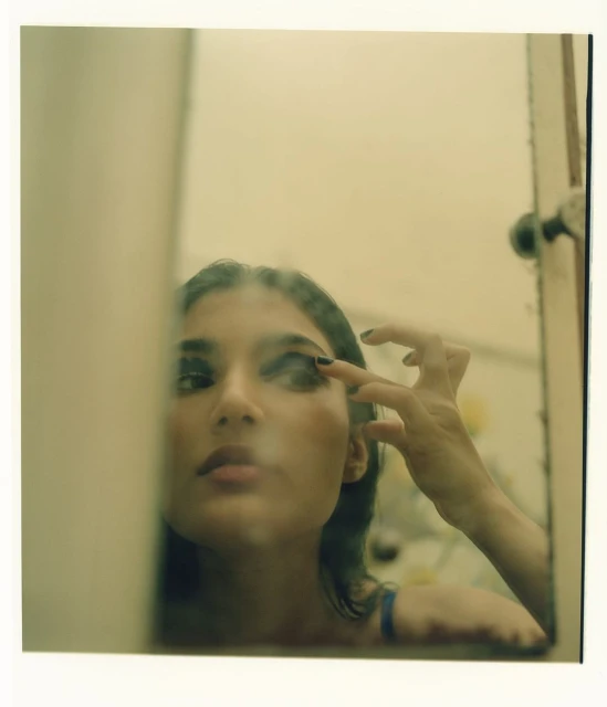 Young woman applying dark eye shadow while looking in a mirror with an off-white frame in an indoor environment.