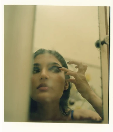 Young woman applying dark eye shadow while looking in a mirror with an off-white frame in an indoor environment.