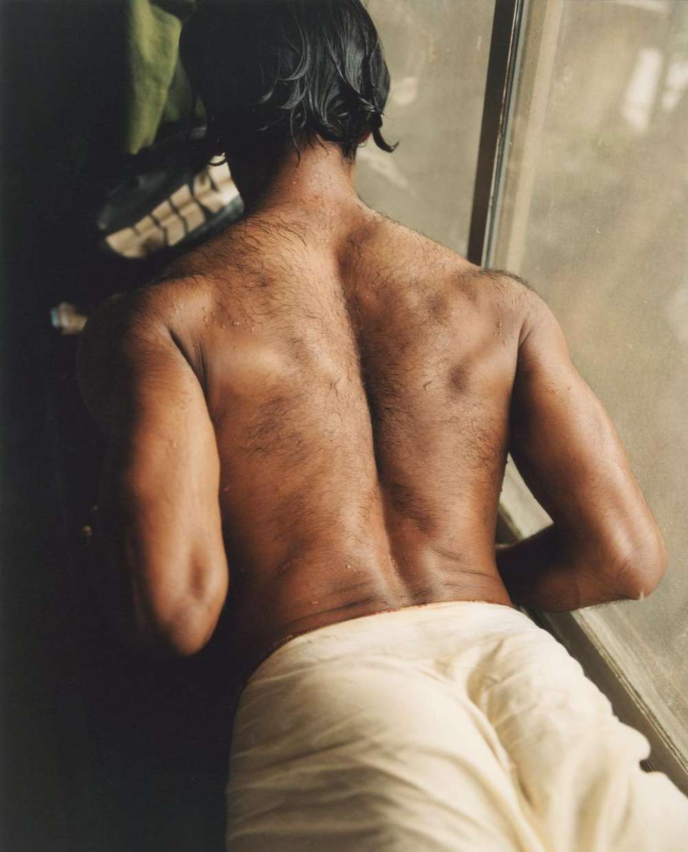 Torso view of adult man with wet black hair, bare back with dark skin, wearing a white towel, looking out window in soft indoor light.