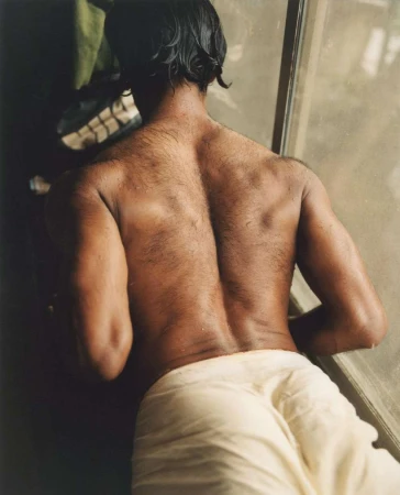 Torso view of adult man with wet black hair, bare back with dark skin, wearing a white towel, looking out window in soft indoor light.