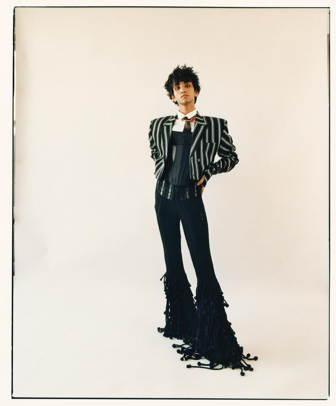 Full-length studio shot of a young adult in a pinstripe jacket, corset, pinstripe pants with tassel details, and a tie against a white backdrop.