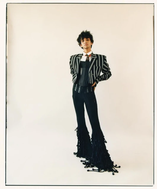 Full-length studio shot of a young adult in a pinstripe jacket, corset, pinstripe pants with tassel details, and a tie against a white backdrop.