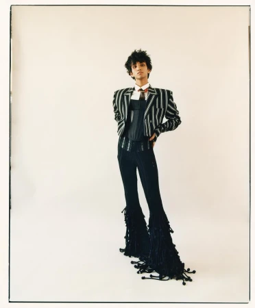 Full-length studio shot of a young adult in a pinstripe jacket, corset, pinstripe pants with tassel details, and a tie against a white backdrop.