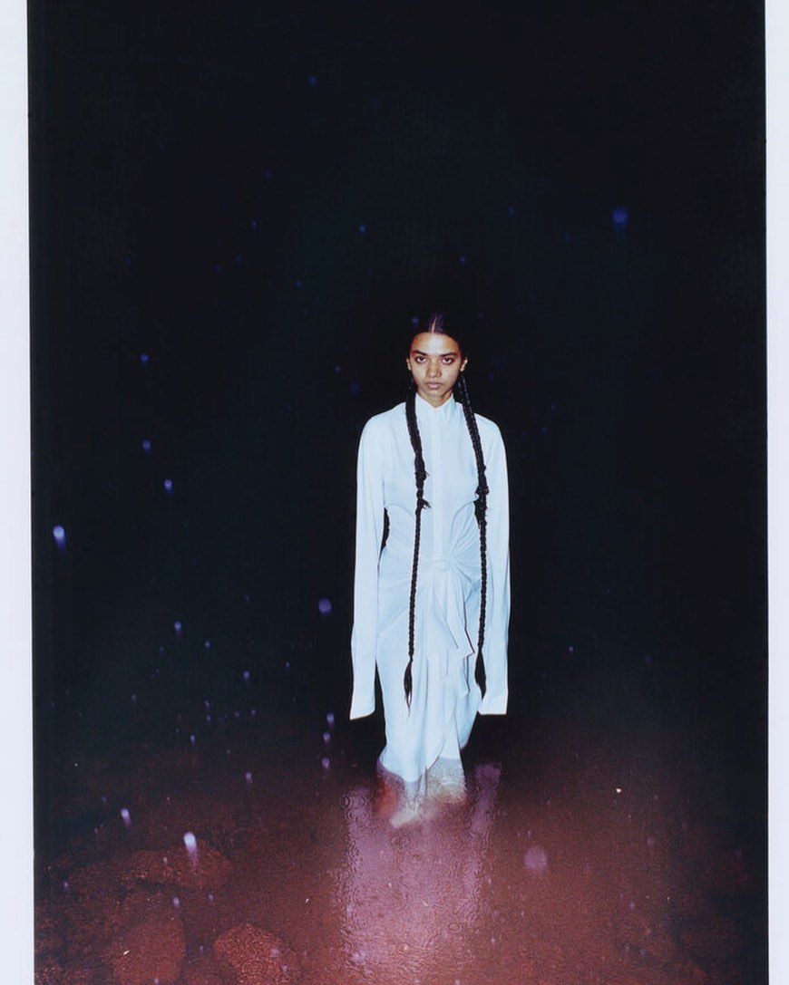 Portrait of a young adult female wading in shallow water against a dark background, wearing a white dress and two long braids.