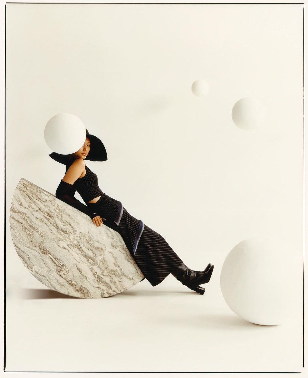 A woman in black outfit and hat poses on a marble semicircle with white spherical props in a fashion photography studio setting.