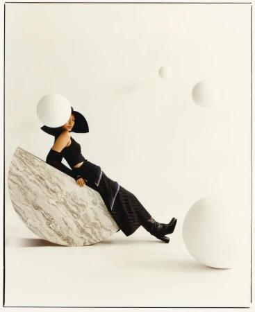 A woman in black outfit and hat poses on a marble semicircle with white spherical props in a fashion photography studio setting.