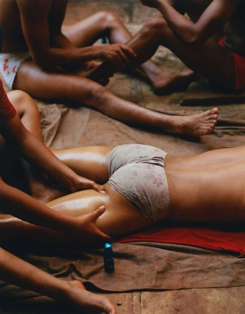 Several individuals are being massaged indoors. The focus is on one woman's lower back and leg being massaged, with oil visible on the skin.