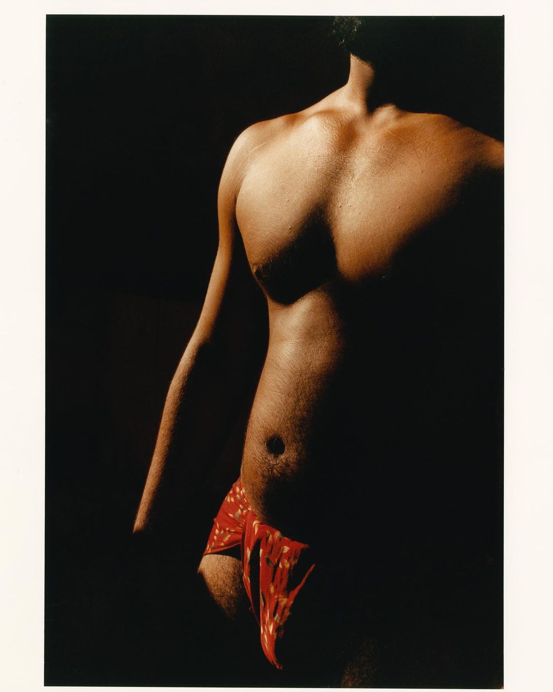 Torso of adult male with dark hair, red patterned fabric around waist, posed in dimly lit studio against dark background.