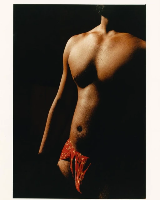 Torso of adult male with dark hair, red patterned fabric around waist, posed in dimly lit studio against dark background.