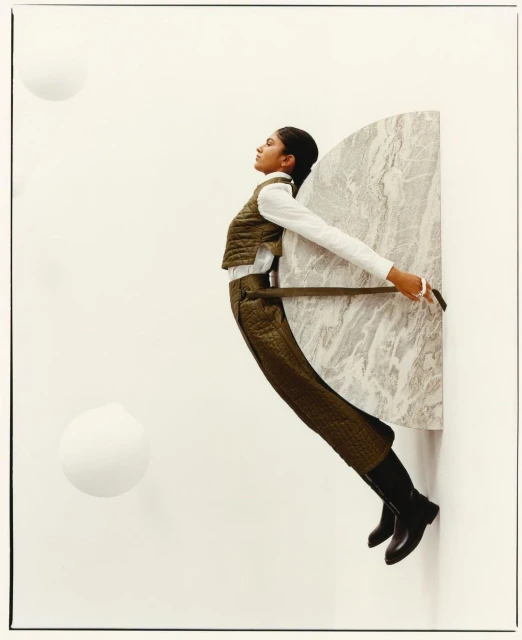 Fashionable woman in olive quilted vest and pants poses holding a marble slab in a studio with white spheres and a white backdrop.