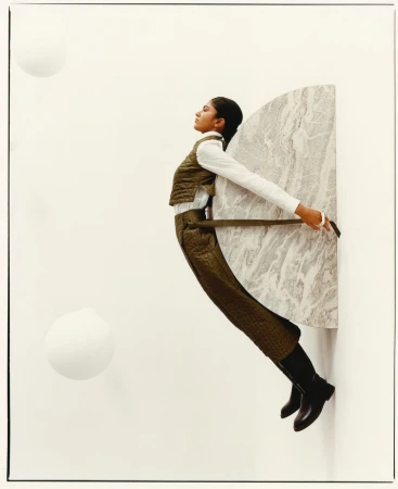 Fashionable woman in olive quilted vest and pants poses holding a marble slab in a studio with white spheres and a white backdrop.