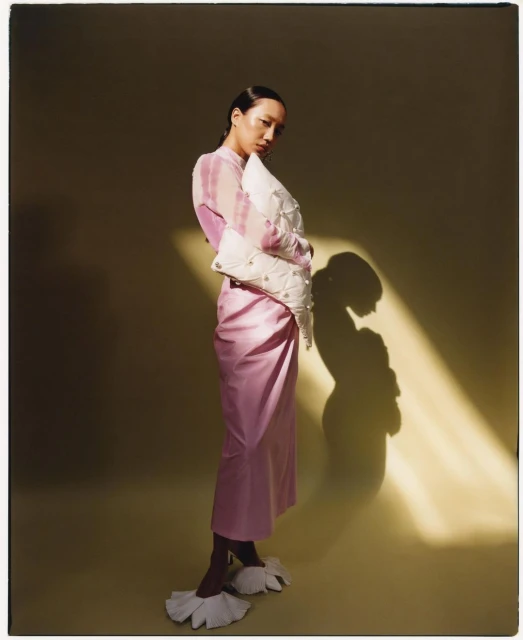 Stylish woman poses in studio wearing pink dress and oversized, pearl-embellished white handbag. Soft light and shadow add depth.