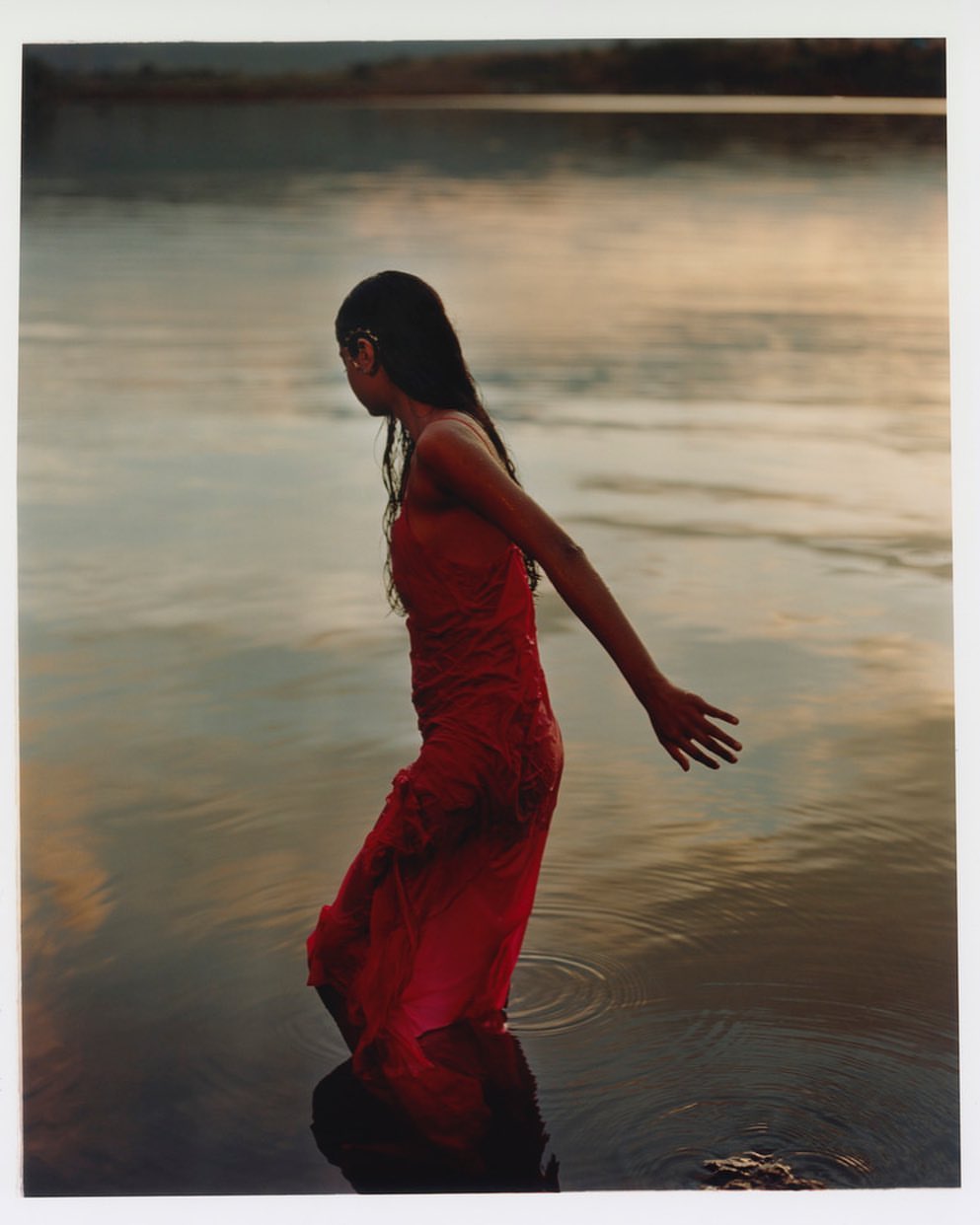 Woman in a red dress walks in lake water at dusk. She is waist-deep and looking away, with hills and sky in the background.