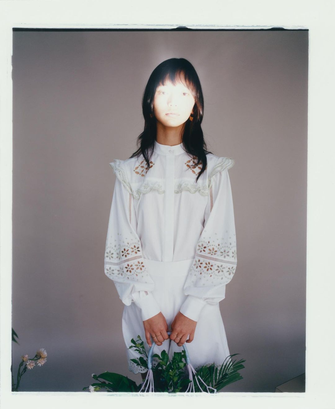 Portrait of a woman in white dress holding a basket of plants in front of a grey background with soft, diffused lighting creating a delicate mood.