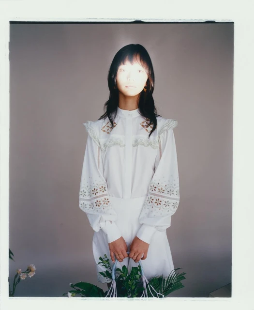 Portrait of a woman in white dress holding a basket of plants in front of a grey background with soft, diffused lighting creating a delicate mood.