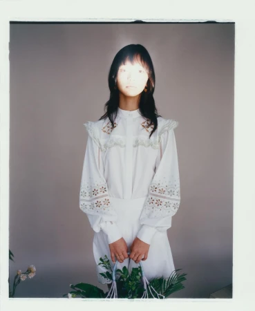 Portrait of a woman in white dress holding a basket of plants in front of a grey background with soft, diffused lighting creating a delicate mood.