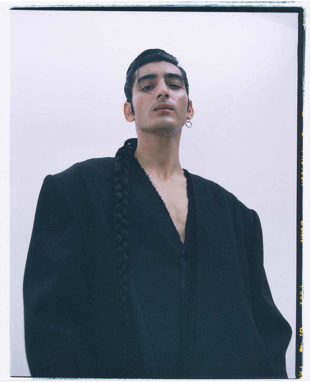 Portrait of a stylish adult male with dark hair, wearing a black oversized jacket. He is posing indoors against a white background.