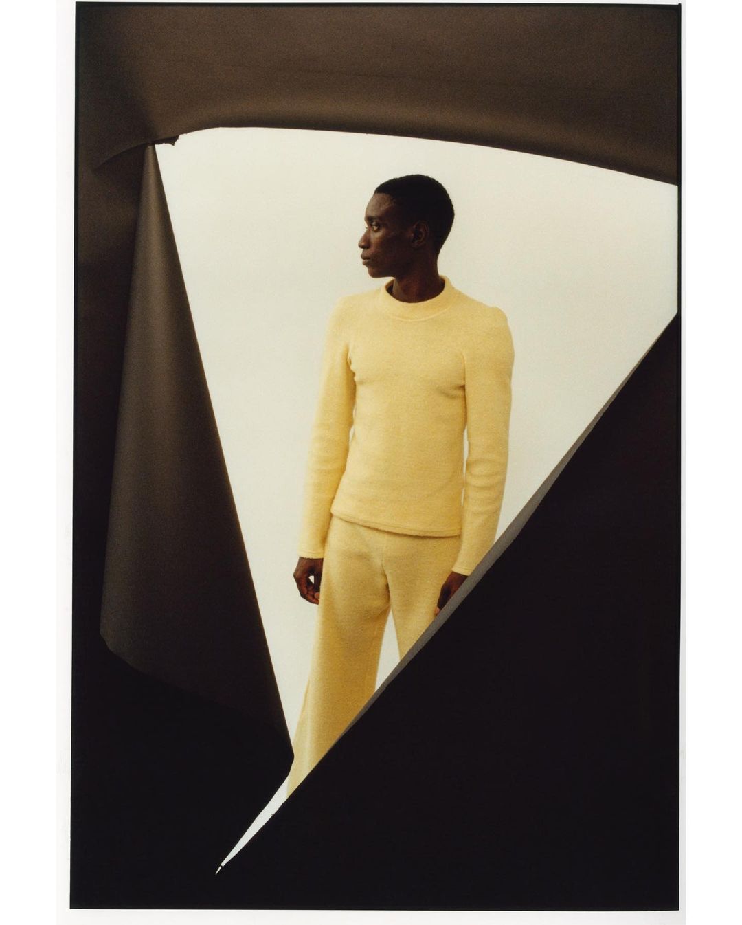 Black man wearing a textured yellow sweater and matching trousers framed by dark abstract paper cutouts in Paris by Rid Burman