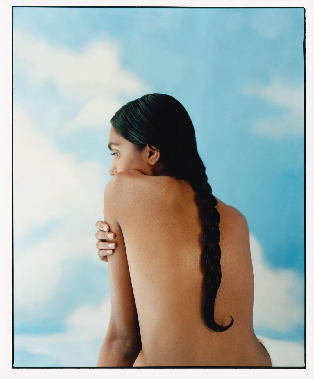Woman with bare back and long braid gazes left in front of a blue sky backdrop with white clouds in a studio setting.