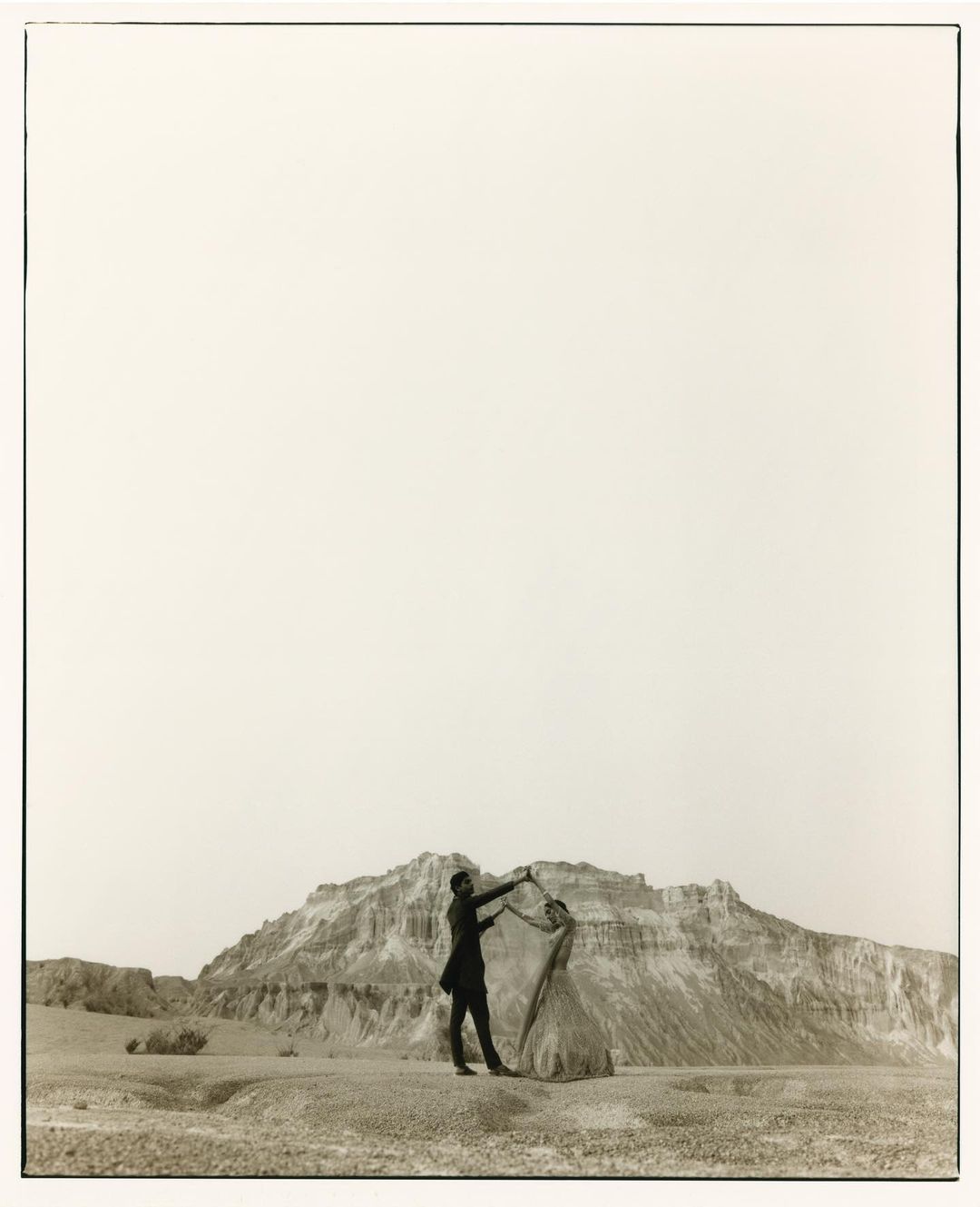 Monochrome image of a couple dancing outdoors in a barren landscape with a rocky mountain backdrop and a vast, bright sky above.