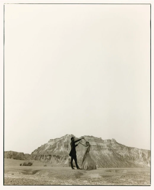Monochrome image of a couple dancing outdoors in a barren landscape with a rocky mountain backdrop and a vast, bright sky above.