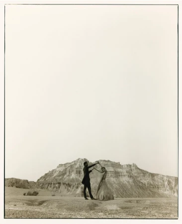 Monochrome image of a couple dancing outdoors in a barren landscape with a rocky mountain backdrop and a vast, bright sky above.