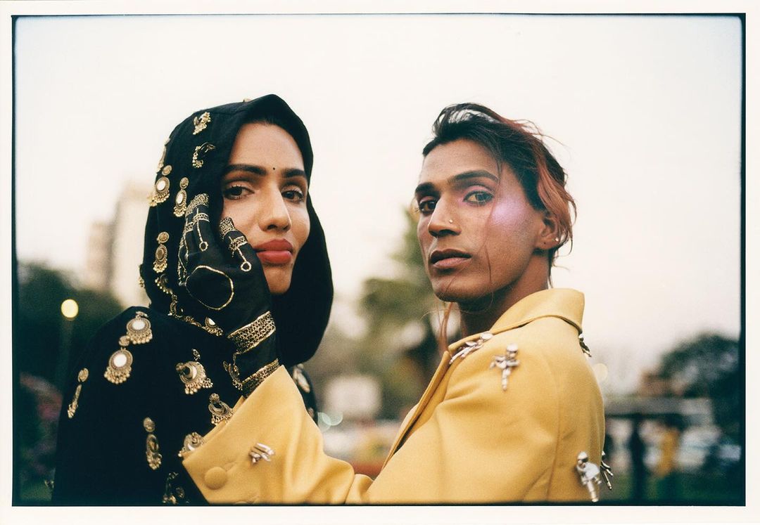 Two fashion models pose outdoors with cityscape. Model in black head covering with ornate gold beading and yellow jacket on model.