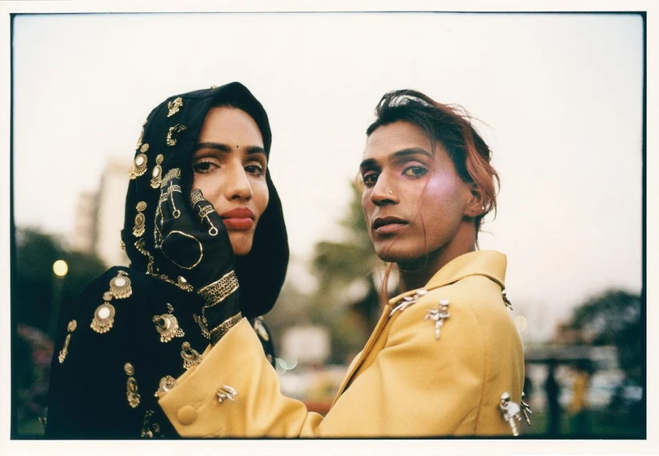 Two fashion models pose outdoors with cityscape. Model in black head covering with ornate gold beading and yellow jacket on model.