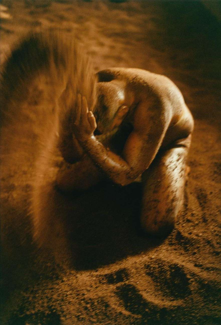 Nude adult kneeling on sand, holding more sand in cupped hands in an outdoor, low-key scene with muted brown and tan tones.
