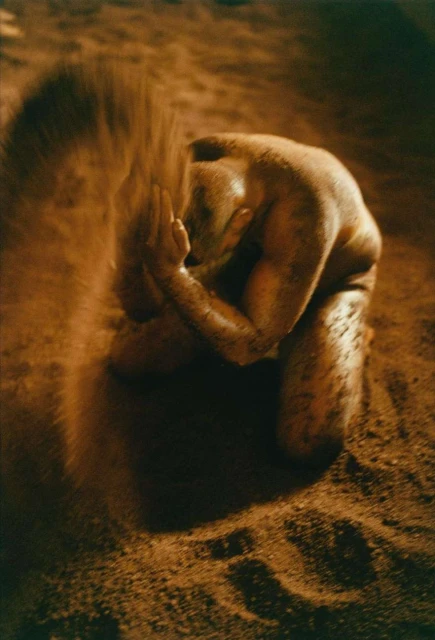 Nude adult kneeling on sand, holding more sand in cupped hands in an outdoor, low-key scene with muted brown and tan tones.