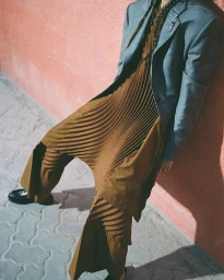 Model wearing a pleated brown dress and grey blazer leaning against a textured peach wall on cobblestone pavement.