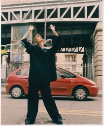 Model in oversized dark suit standing on a city street with arms raised as giant bubbles float past, with a red car parked in the background