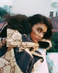 Close-up portrait of a person with long dark hair leaning over a white architectural railing, wearing a patterned jacket.