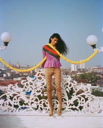 Model with long dark hair wearing a purple top and patterned tights standing on a white rooftop against a city skyline.