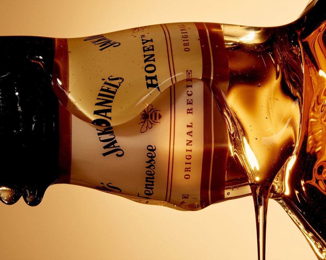 A close-up photograph of a Jack Daniel's Tennessee Honey bottle with honey dripping down the side against a neutral background.