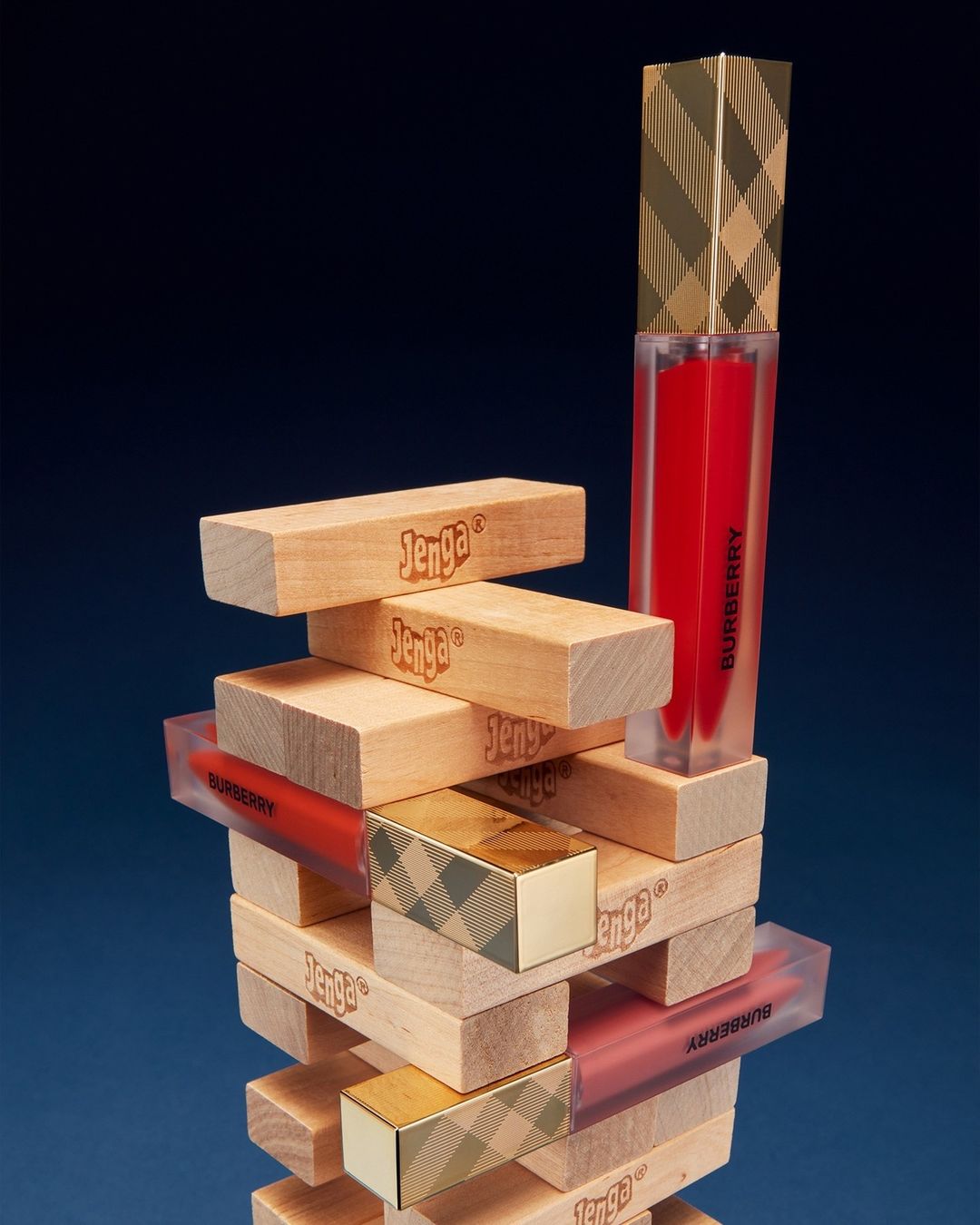 Burberry lip gloss tubes stacked on a Jenga game tower against a dark blue background, showing beauty and games.